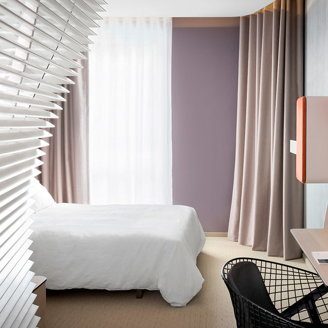 Camera d'albergo moderna con letto bianco, parete color malva, tende tortora, veneziane bianche e pannello lampada coralline.