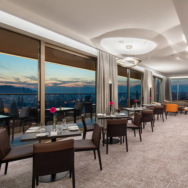 Elegante ristorante con vista panoramica sulla città al tramonto, con lampada a sospensione circolare e illuminazione indiretta calda sopra i tavoli.
