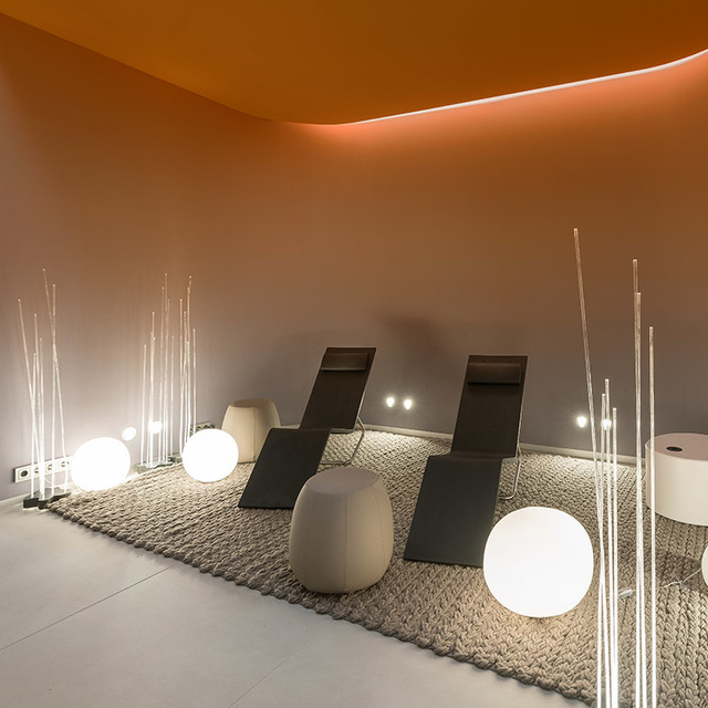 Area relax con lampade da terra sferiche bianche luminose e bastoni luminosi alti su tappeto, accanto a sedie reclinabili scure sotto luce soffitto arancione