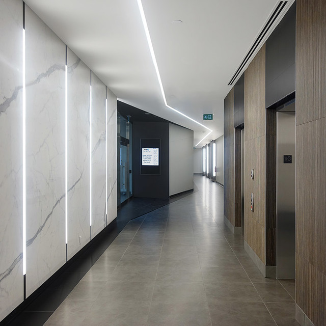 Corridoio moderno con luci LED lineari a soffitto e strisce luminose verticali a parete che illuminano pannelli in marmo vicino agli ascensori.