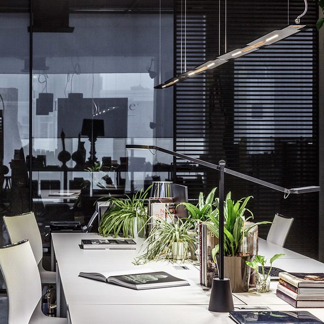 Modern office workspace with a sleek black linear pendant lamp overhead and an adjustable desk lamp, surrounded by green plants on a white desk.