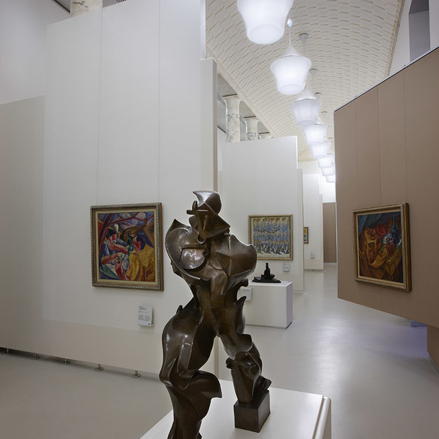 Museum gallery with bronze sculpture, colorful paintings on white walls, lit by spherical pendant lamps with warm white light.