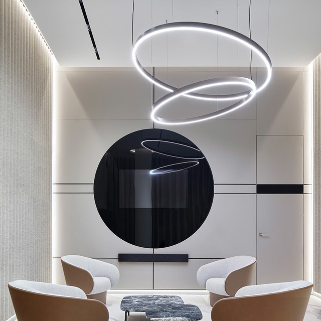 Salotto moderno con anelli a sospensione LED circolari sovrapposti in nero, sospesi sopra area salotto con grande arte murale circolare nera.