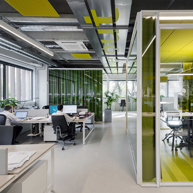 Ufficio open space moderno con luci lineari a soffitto, sala riunioni con pareti in vetro, accenti verdi e dipendenti al lavoro a scrivanie bianche