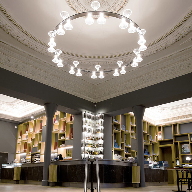 Circular chandelier with multiple bell-shaped glass diffusers illuminating an elegant retail space with ornate plaster ceiling moldings.