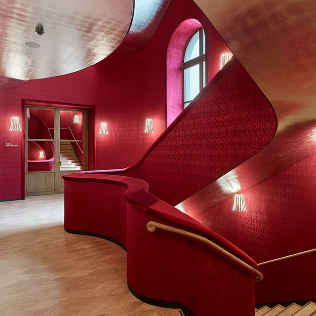 Curved red staircase with patterned walls, illuminated by Artemide wall sconces with white fringe shades casting warm light.