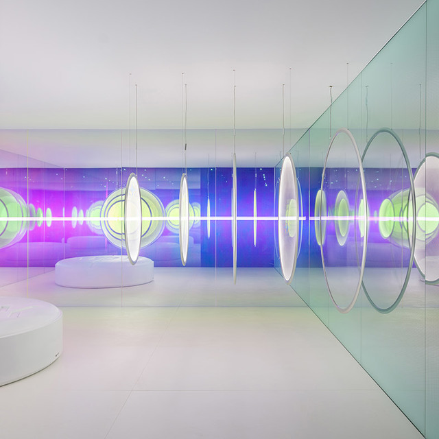 Galleria bianca futuristica con lampade a sospensione ovali Artemide su sottili cavi, riflesse in pareti di vetro tra proiezioni luminose viola e verde