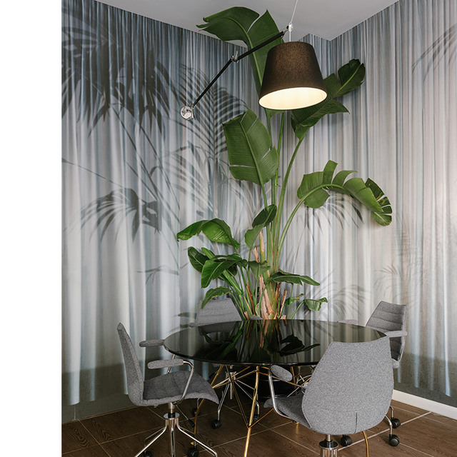 Floor lamp with dark conical shade on curved arm over a round black table with chairs, surrounded by tropical plants and sheer curtains