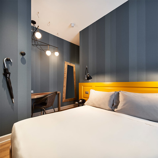 Hotel bedroom with blue striped walls, yellow headboard, pendant lamp with two bulbs, and a black adjustable wall lamp above the desk.