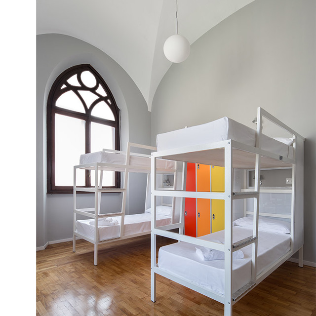 Hostel dormitory with white bunk beds under vaulted ceiling, Gothic window, colorful lockers, and a spherical white pendant lamp.