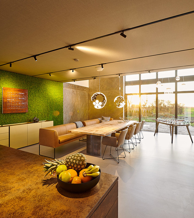 Zona pranzo open space moderna con lampade a sospensione Artemide sopra un lungo tavolo, binari luminosi, parete di muschio verde e finestre a tutta altezza