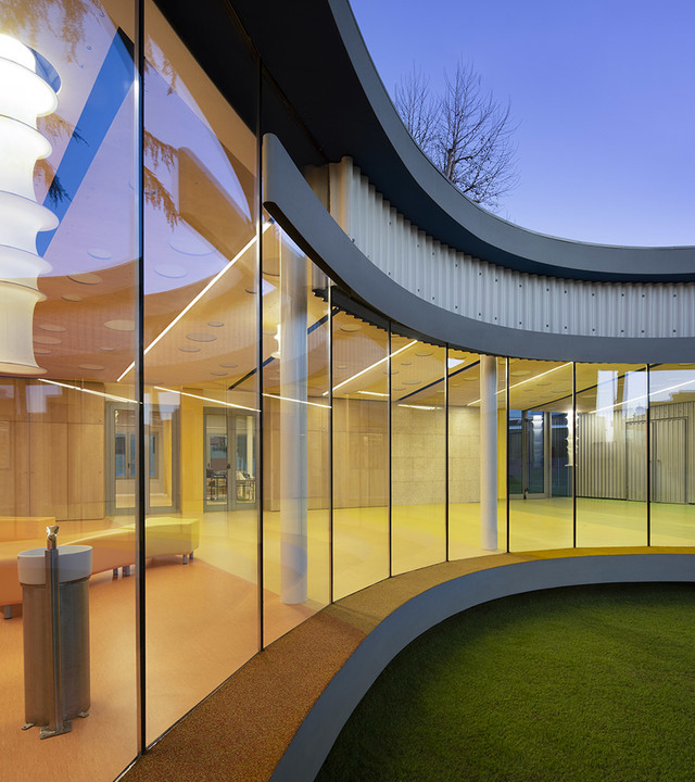 Facciata curva in vetro di un edificio moderno al crepuscolo, interno illuminato da lampade lineari LED Artemide a soffitto con luce d'ambiente calda.