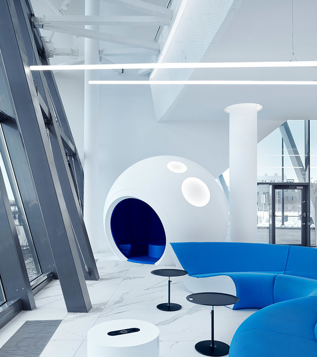 Lounge ufficio moderno con lampade lineari a soffitto Artemide, area seduta a capsula sferica bianca e vivaci divani modulari blu su pavimento in marmo