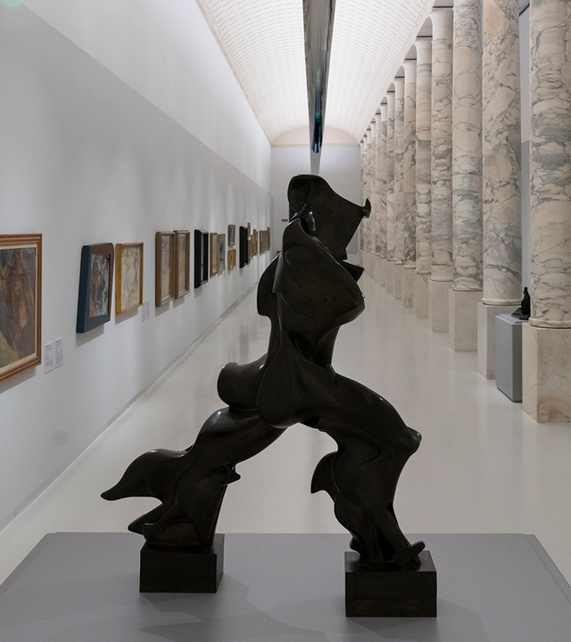 Galleria museale con grande scultura astratta in bronzo scuro, dipinti su pareti bianche e colonne in marmo sotto illuminazione lineare a soffitto.