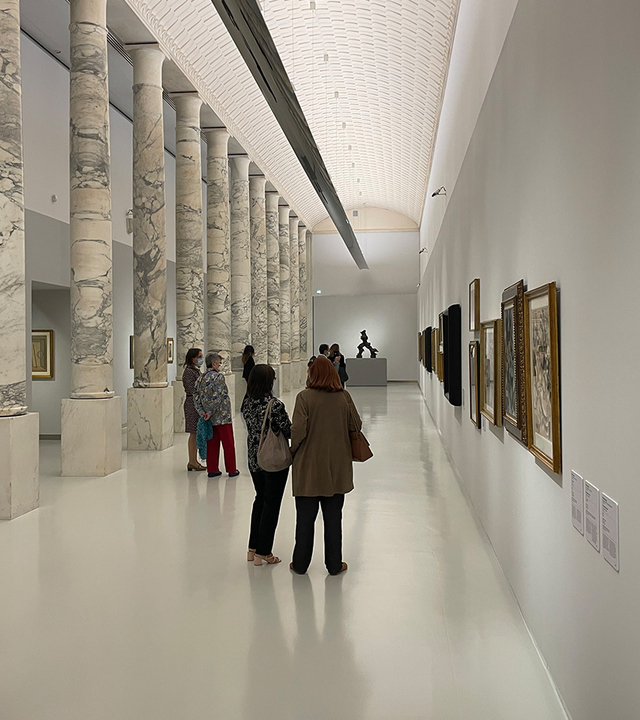Galleria museale con colonne in marmo, visitatori che osservano opere d'arte su pareti bianche, illuminata da soffice luce indiretta a soffitto.