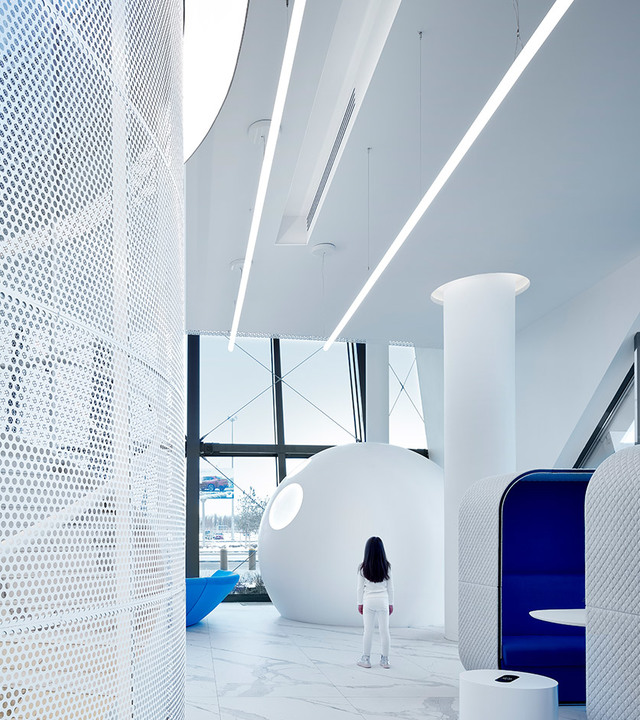 Interno bianco futuristico con luci lineari a soffitto, pannello forato, struttura sferica a baccello e una persona in piedi nello spazio