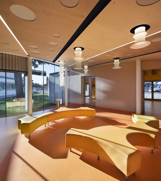 Lounge moderno con sedute curve gialle, pavimento terracotta caldo e lampade a sospensione a livelli bianchi in uno spazio luminoso