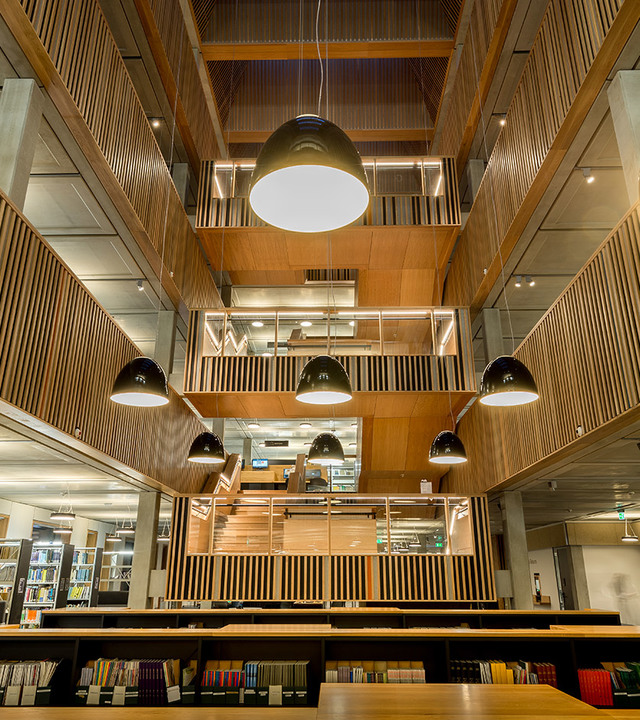 Multi-level library atrium with black dome pendant lights at various heights, warm wood paneling, and a large circular ceiling lamp above.