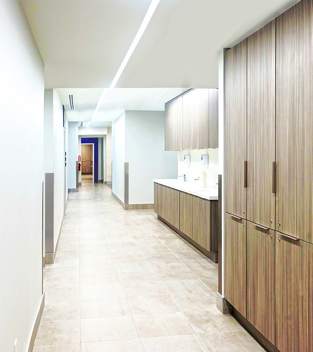 Corridoio ufficio moderno con illuminazione LED lineare a incasso su soffitto bianco, mobili con finitura legno e pavimento in piastrelle neutre.