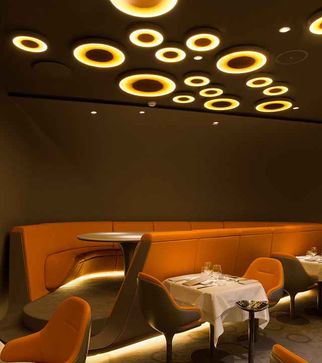 Interno di ristorante con luci a soffitto circolari Artemide in caldi toni dorati sopra sedute a divano arancioni e tavoli da pranzo con tovaglia bianca.
