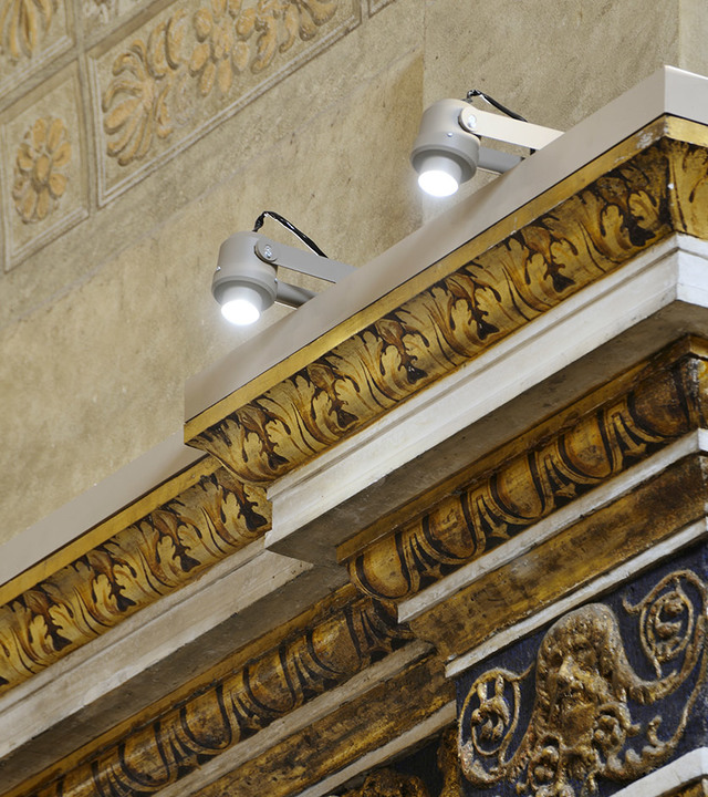 Two Artemide adjustable outdoor spotlights mounted on ornate gilded cornice of a historic building, illuminating architectural details.