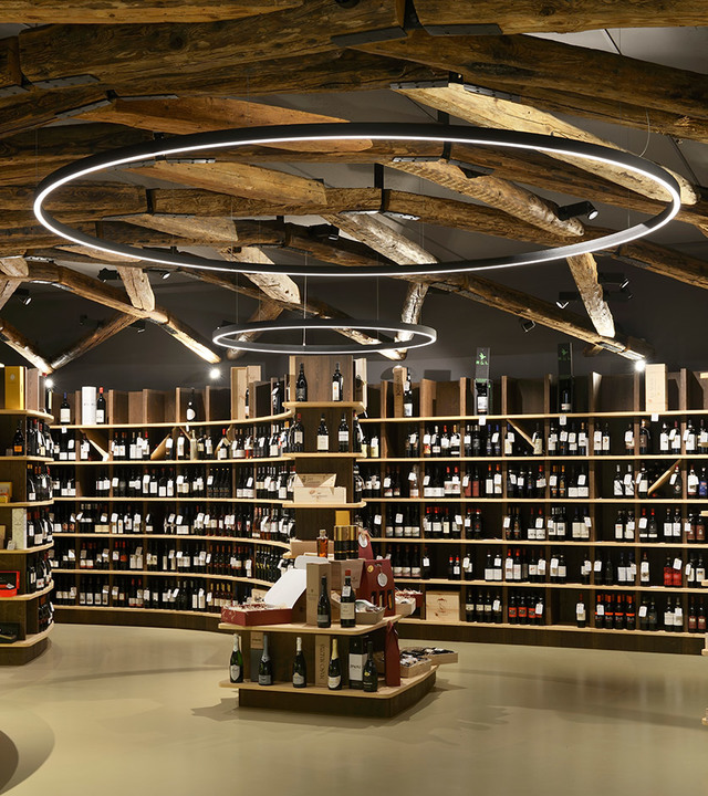 Interno di enoteca con anelli a sospensione LED circolari Artemide che illuminano soffitto con travi in legno rustico e scaffali di bottiglie di vino