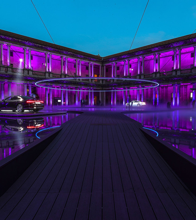 Cortile neoclassico immerso in luce viola con installazione ad anello neon luminoso, pavimento riflettente e due auto di lusso in esposizione