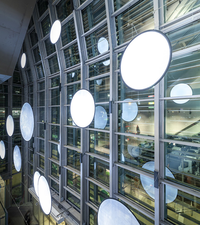 Multiple large circular LED pendant lights suspended at varying heights in front of a multi-story glass curtain wall facade.