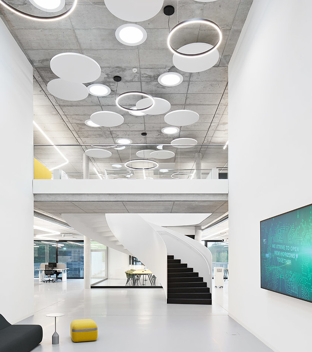 Atrio di ufficio moderno con soffitto in cemento a vista, lampade a sospensione circolari e ovali Artemide, scala a chiocciola e soppalco aperto.