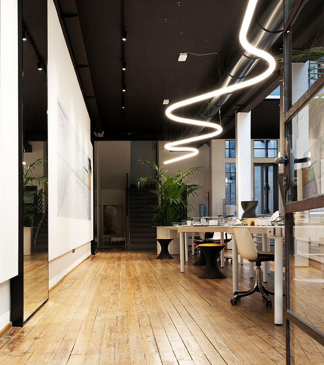 Lampada a sospensione Artemide Alphabet of Light a forma di onda sinuosa illumina un ufficio moderno con pavimento in legno e soffitto scuro.