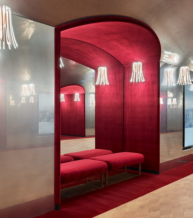 Elegant red-upholstered alcove with arched mirror and Artemide wall lamps with white fringe shades creating warm ambient light.