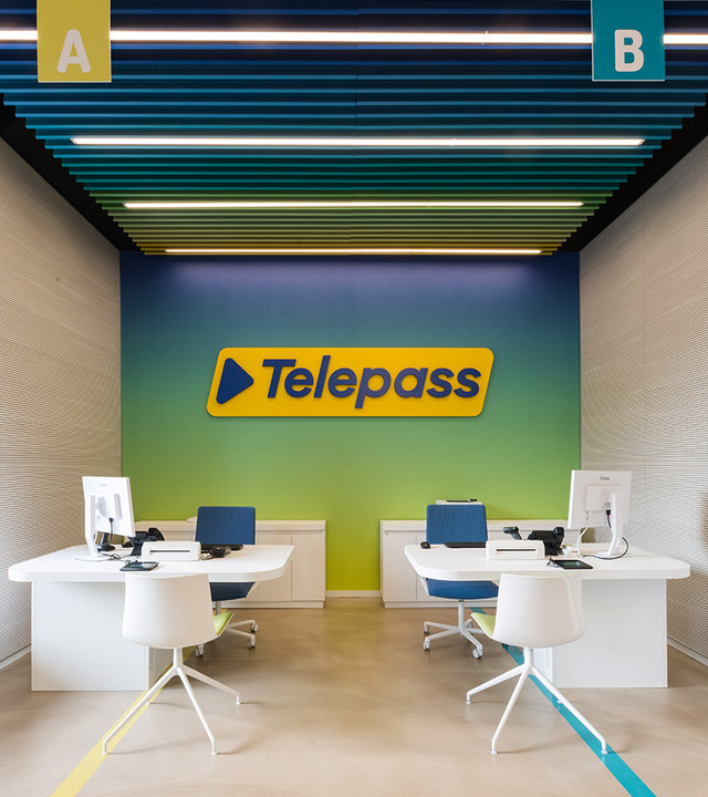 Ufficio Telepass con due postazioni di lavoro sotto lampade lineari da soffitto Artemide su un soffitto a listelli colorati verde acqua e giallo.