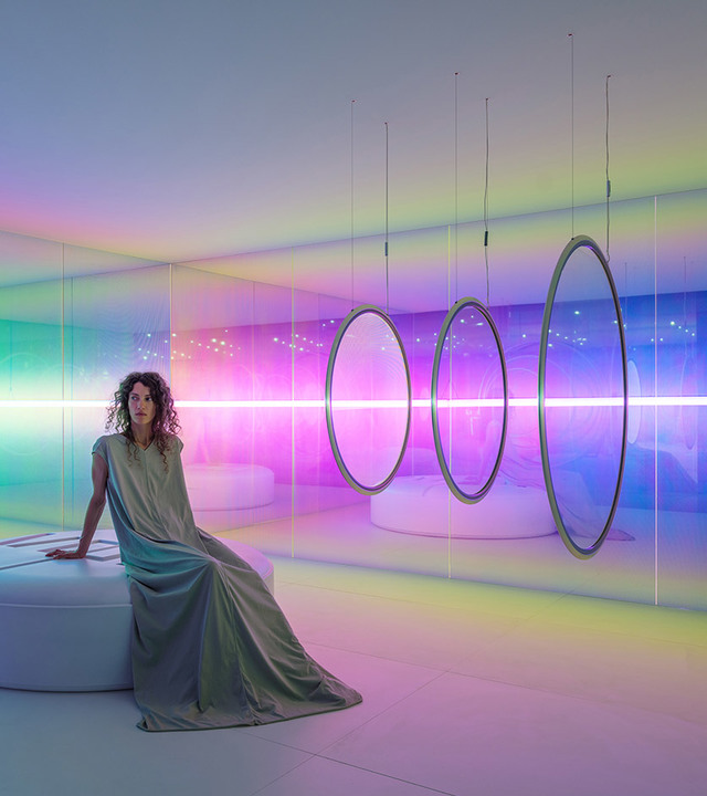 Donna in abito drappeggiato seduta in una stanza immersiva con illuminazione RGB colorata, anelli a sospensione circolari e vividi fasci di luce rosa e verde