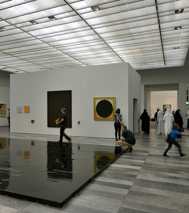 Modern art museum interior with recessed ceiling lighting illuminating visitors viewing abstract paintings on white walls.