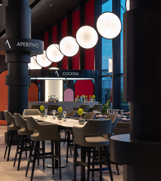 Area bar di un ristorante con una fila di sei lampade a sospensione Artemide a globo bianco sferico sopra tavoli alti e sgabelli grigi.