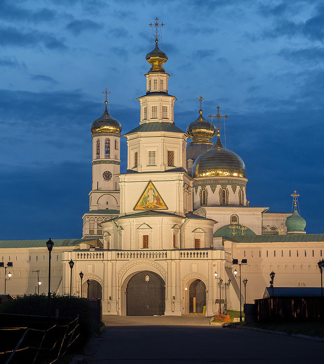 Russian Orthodox monastery illuminated at dusk, white facade with golden and green onion domes glowing under a dramatic blue sky