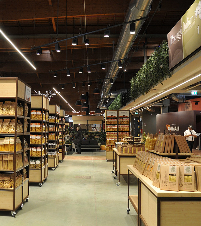 Retail food store interior with linear LED ceiling lights and recessed spotlights illuminating pasta and dry goods shelves.