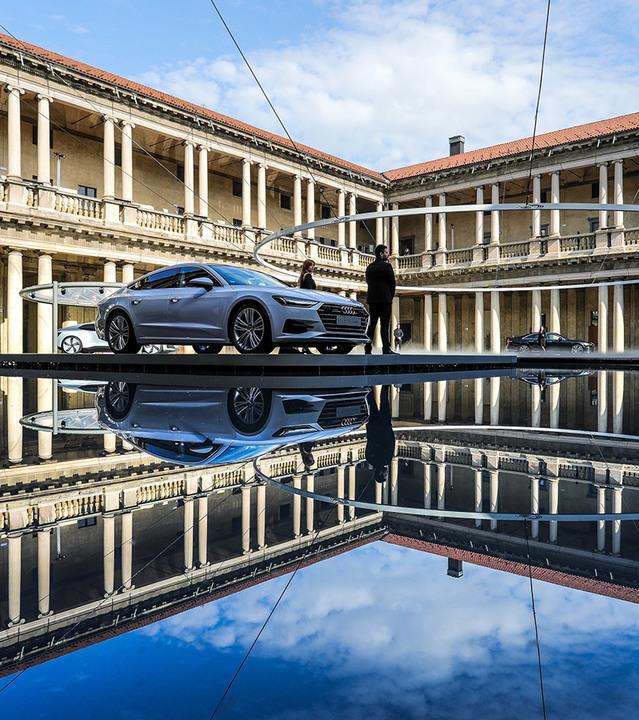 Berlina Audi esposta su una piattaforma rialzata in un cortile colonnato classico, riflessa in una superficie d'acqua a specchio.