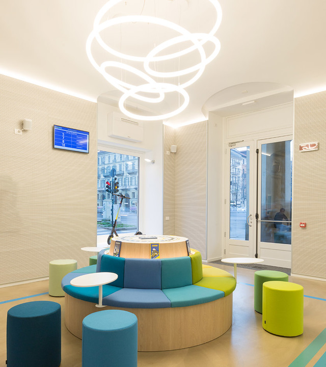 Sala d'attesa moderna con seduta circolare e sgabelli colorati, illuminata dalla lampada a sospensione Artemide Overlap con anelli luminosi intrecciati.