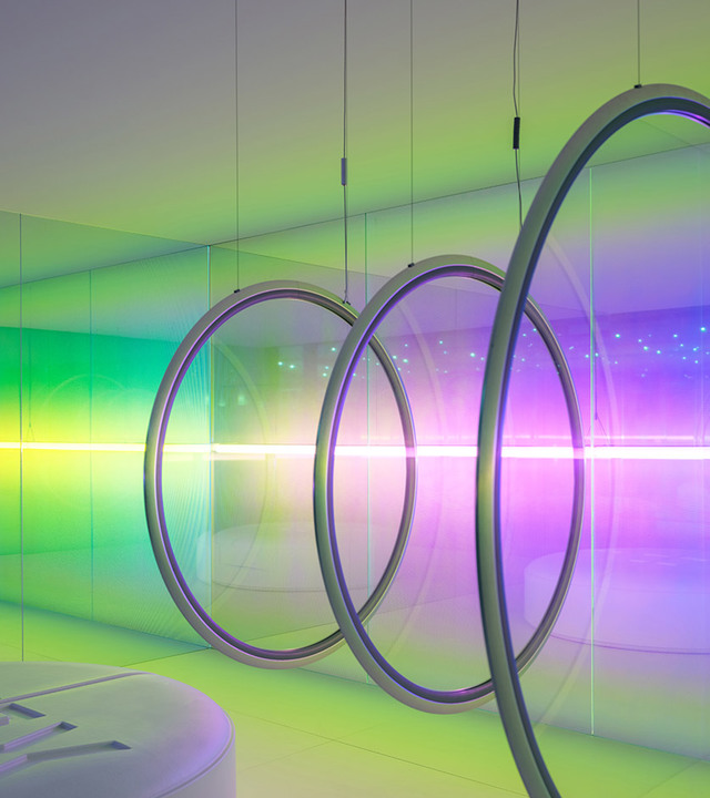 Grandi anelli circolari a sospensione emettono vivace luce verde, viola e bianca, creando un'atmosfera cromatica immersiva in una stanza a specchi.