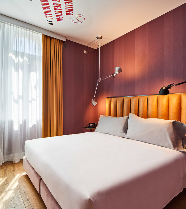 Hotel bedroom with double bed, burgundy walls, and two Artemide Tolomeo wall lamps with adjustable arms on each side.