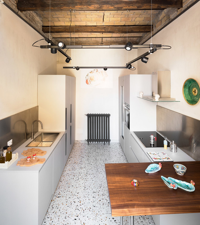 Cucina moderna con sistema di illuminazione a binario Artemide con faretti neri orientabili sospesi da travi in legno a vista su pavimento in terrazzo.