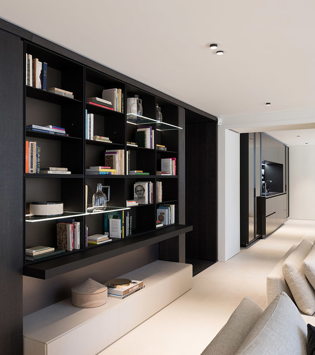 Soggiorno moderno con libreria a muro scura illuminata da strisce LED integrate, faretti a soffitto e divano in toni chiari vicino.
