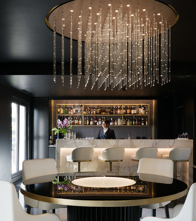 Elegant bar lounge with a large circular chandelier featuring cascading crystal rods over a round black and gold table with white chairs.