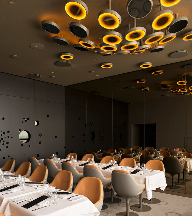 Ristorante elegante con lampade a sospensione circolari Artemide che emanano luce ambrata su soffitto scuro, sopra tavoli apparecchiati e sedie rivestite in cuoio.