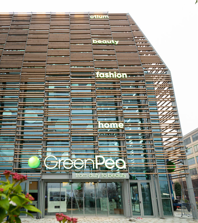 GreenPea sustainable shopping center in Turin, Italy, featuring a modern facade with wooden slats and glass, displaying category signs.