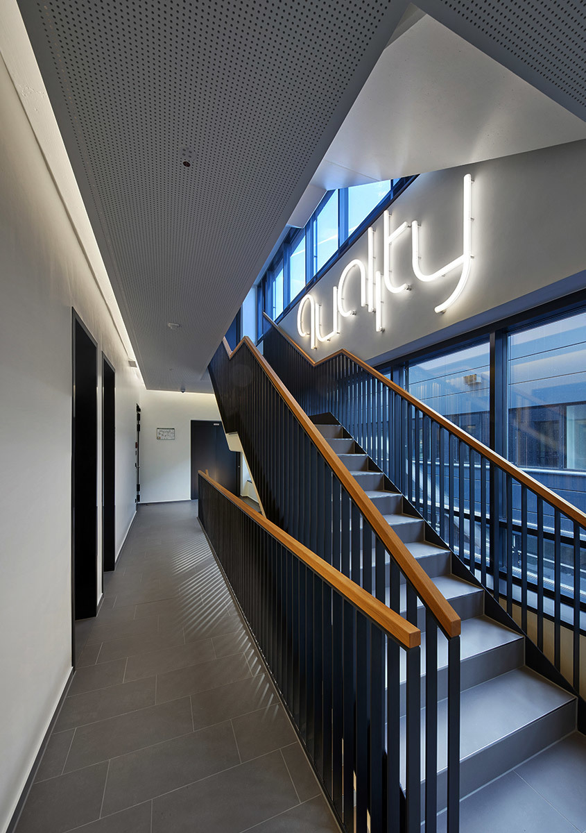 Vano scala di un ufficio moderno con ringhiere in metallo scuro, corrimano in legno e un'insegna al neon luminosa 'quality' sulla parete.