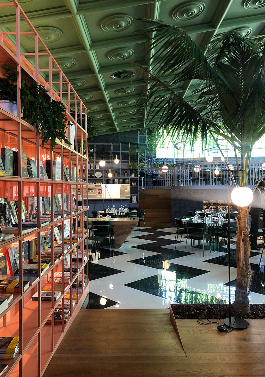 Restaurant interior with Artemide floor lamps, pink metal shelving, tropical palms, checkered floor, and ornate green ceiling.