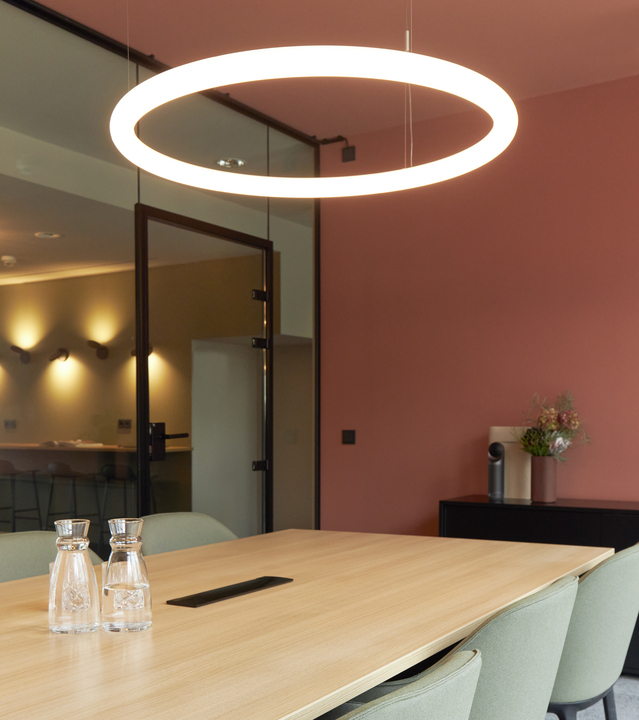 Artemide circular LED pendant lamp suspended over a wooden conference table in a modern meeting room with pink walls.