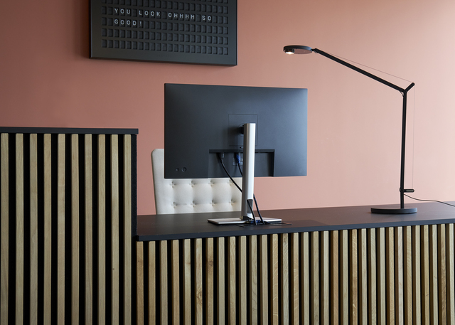 Artemide dark desk lamp with adjustable arm on a modern reception desk with slatted wood panels and a monitor.